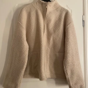 Beige teddyjacka från Amisu - Mysig beige teddyjacka pile, från Amisu med dragkedja framtill. Jackan har en hög krage och långa ärmar, perfekt för kyligare dagar. Den är tillverkad i ett mjukt och fluffigt material som ger en skön känsla.