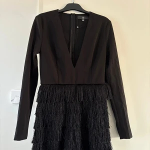 Svart klänning med fransar - Snygg svart klänning med långa ärmar och djup v-ringning. Kjoldelen har trendiga fransdetaljer som ger en cool look. Perfekt för en kväll ute eller en speciell tillställning. Storlek S
