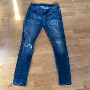 Blå jeans med slitningar - Säljer ett par blå jeans med slitningar på knäna och en snygg tvätt. De har en klassisk femficksdesign och en läderpatch på baksidan av midjan. Perfekta för en avslappnad stil.