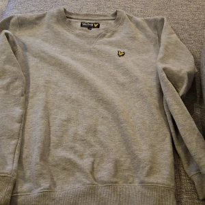 Grå tröja från Lyle & Scott 14-15 år - Säljer en stilren grå tröja från Lyle & Scott med deras ikoniska logga på bröstet. Tröjan har en klassisk passform med ribbade muddar vid ärmslut och nederkant. 