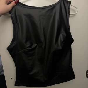 Ny svart ärmlös topp i skinn - Helt ny svart ärmlös topp i glansigt skinn matrial och aldrig använd. Perfekt för en cool och edgy look. Passar bra till både jeans och kjol.