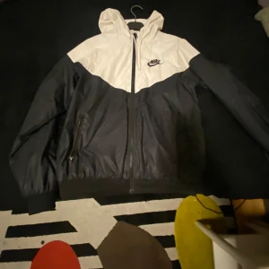 Svart och vit vindjacka från Nike vår/sommar.  - Säljer en snygg svart och vit vindjacka från Nike. Jackan har en dragkedja framtill och en praktisk huva. Perfekt för blåsiga dagar och har en sportig look. Den är tillverkad i ett lätt syntetmaterial.