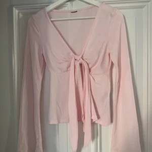 Ljusrosa långärmad topp med knyt - Säljer en söt ljusrosa topp med långa ärmar och knyt framtill. Toppen har en v-ringad design som ger en feminin touch. Perfekt för att lägga till en romantisk känsla till din outfit.