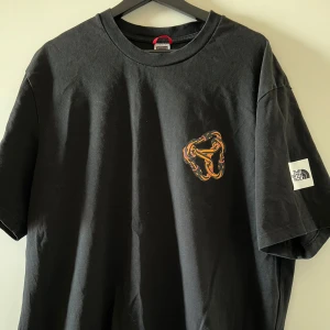Svart t-shirt från The North Face - Säljer en svart t-shirt från The North Face med ett coolt grafiskt tryck i orange och rosa på både fram- och baksidan. T-shirten har korta ärmar och en normal passform. Perfekt för en avslappnad stil!