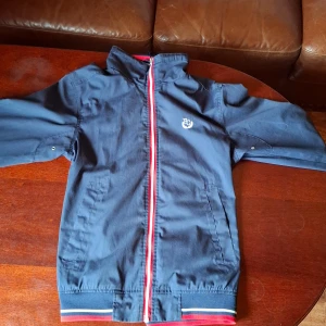 Blå jacka från Etirel - Säljer en snygg blå jacka från Etirel med dragkedja och broderad logga på bröstet. Jackan har en rutig insida och ribbade muddar. Perfekt för en sportig look!