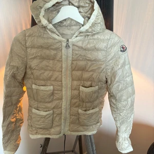 Moncler jacka  - Snygg beige dunjacka från Moncler med huva och dragkedja framtill. Jackan har två praktiska fickor och Moncler-logga på ärmen. Perfekt för kyligare dagar. Självklart äkta och köptes på NK
