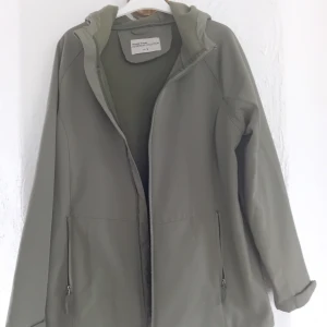 Grön vindjacka från My Ware - Säljer en stilren grön vindjacka från My ware i storlek L. Jackan har en praktisk huva och dragkedja framtill. Lite softshelltyg och nägut fodrad.Perfekt för vårpromenaden!