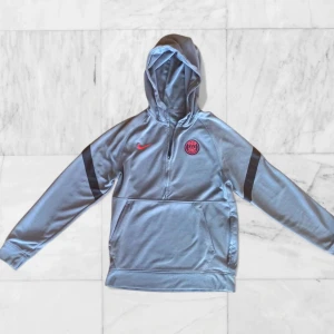 Grå hoodie från Nike med PSG-logga - PSG quarter zip hoodie från Nike, storlek S. Har inte använt den på ett tag, den har bara legat i garderoben. Är fortfarande i bra skick överlag. Det finns en liten blå fläck som kanske går bort i tvätten, och Nike-sömmen på loggan är något sned, men inget stort eller synligt när den bärs. Superkomfortabel och lätt att styla, perfekt för fans eller casual looks. Modellen är 170 cm lång och väger 60 kg. Passar bra 