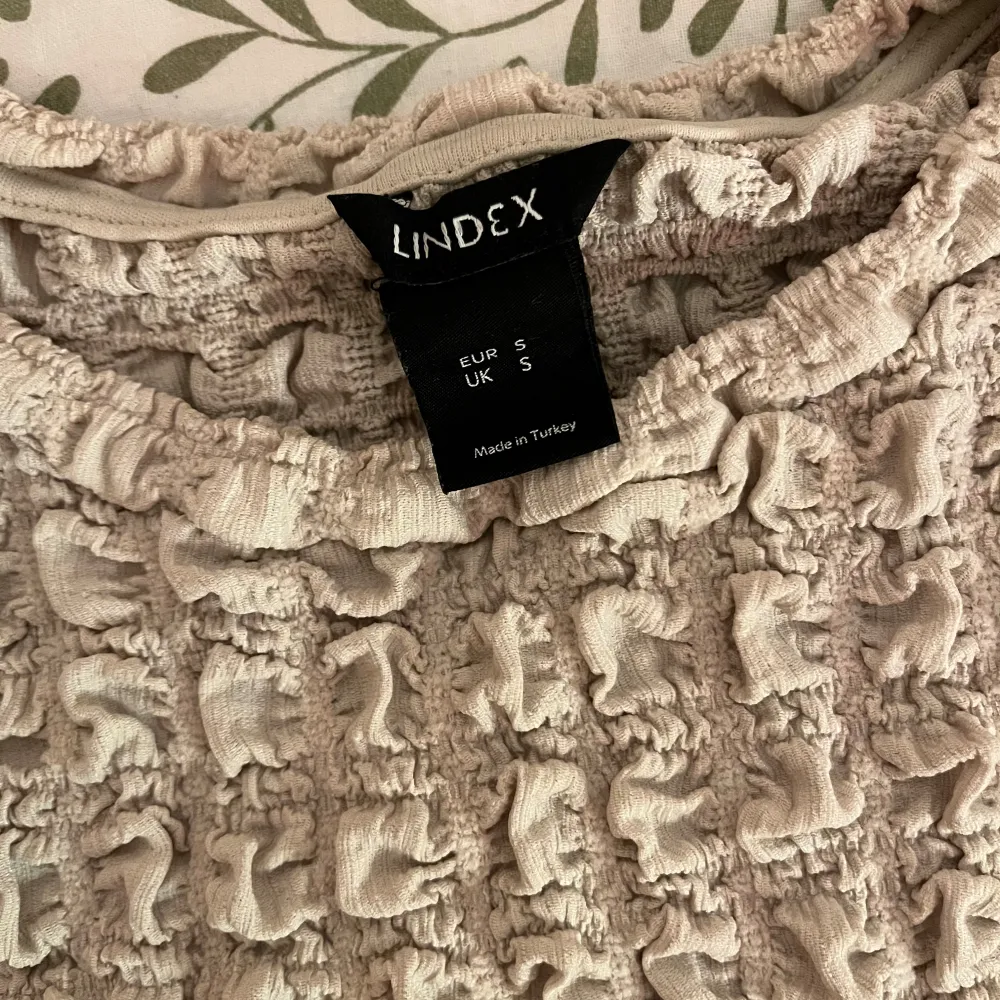 Säljer en unik beige topp från Lindex med ett texturerat, rynkat mönster. Toppen har korta ärmar och en rund halsringning. Perfekt för att ge din outfit en intressant touch. 🌟. Neuletakit & Villapaidat.