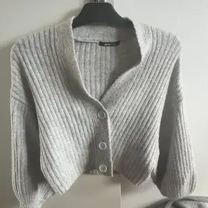 Säljer en stilren grå ribbad kofta från Gina. Koftan har en v-ringad design med tre stora knappar framtill och långa ärmar. Perfekt för kyliga kvällar.