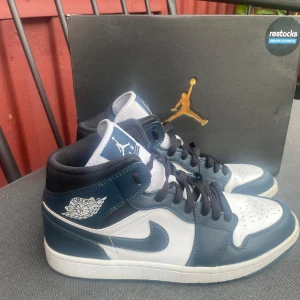 Nike Air Jordans 1 ”Dark teal” mid - Snygga Nike Air Jordans i en stilren blå och vit färgkombination. Skorna har klassisk snörning och den ikoniska Air Jordan-loggan på sidan. Perfekta för dig som vill ha en sportig och trendig look nu inför sommaren.