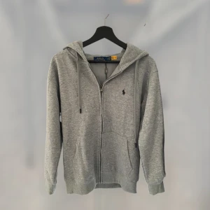 oanvänd zip hoodie från Ralph Lauren - Säljer en stilren grå hoodie från Polo Ralph Lauren med dragkedja och det klassiska logotypbroderiet på bröstet. Perfekt för en avslappnad look. Den har en justerbar huva och ribbade muddar.