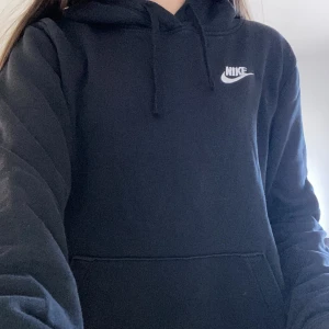 Svart hoodie från Nike - Säljer en klassisk svart hoodie från Nike med vit logga på bröstet. Tröjan har en stor ficka framtill och justerbar huva med dragsko. Perfekt för en avslappnad stil.