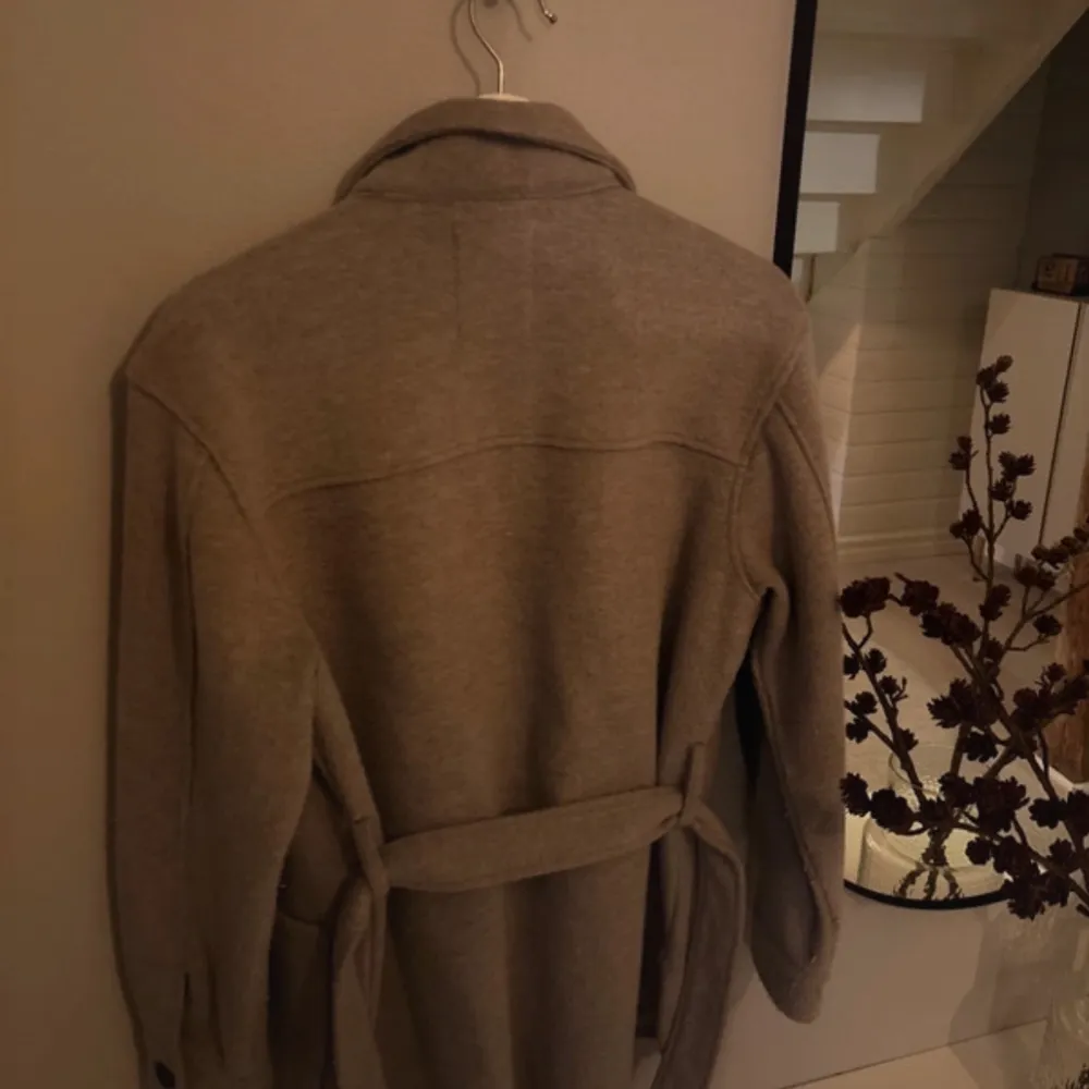 Säljer en stilren beige kappa från Dobber. Den har en klassisk krage och knyts med ett bälte i midjan. Perfekt för höst och vår. Kappan har långa ärmar och två stora fickor framtill.. Takit.