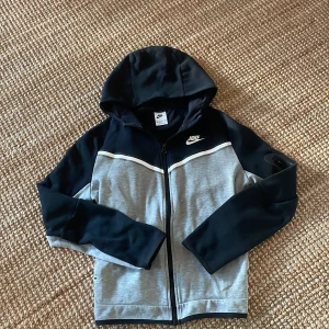 Grå och svart nike tech - Säljer en snygg grå och svart hoodie från Nike med dragkedja och huva då sen inte längre passar mig. strlk är 158-170cm. Jackan har en cool ficka med dragkedja på ärmen och Nike-loggan på bröstet. 