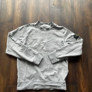 Grå sweatshirt från Stone Island - Säljer en stilren grå sweatshirt från Stone Island. Tröjan har långa ärmar och en rund halsringning. Den ikoniska loggan sitter på ärmen. Perfekt för en avslappnad stil.