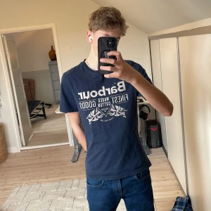 Marinblå t-shirt från Barbour - Säljer en blå t-shirt från Barbour med tryck på framsidan. T-shirten är kortärmad och tillverkad i bomull. Perfekt för en avslappnad stil.pris kan diskuteras