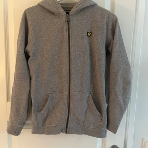 Grå hoodie från Lyle & Scott - Säljer en stilren grå hoodie från Lyle & Scott med dragkedja och huva. Tröjan har två praktiska fickor framtill och ett litet gult emblem på bröstet. Perfekt för en avslappnad stil. Bara använt max tre gånger. 