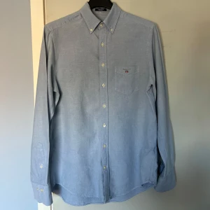 Blå Oxfordskjorta från GANT - Säljer en klassisk blå Oxfordskjorta från GANT med normal passform. Skjortan har en bröstficka med broderad logotyp och knappar framtill. Perfekt för en stilren look.