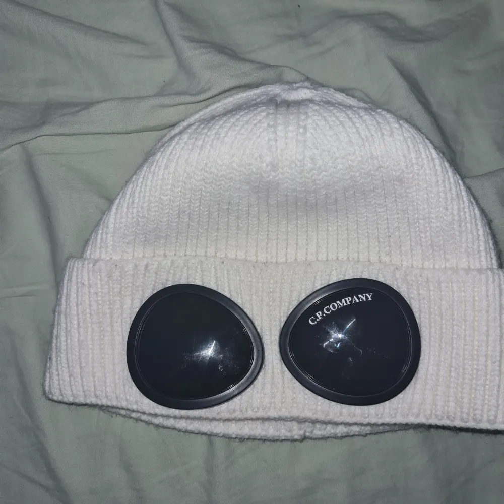 Snygg vit mössa från C.P. Company med unika glasögondetaljer på framsidan. Tillverkad i 100% bomull för en bekväm passform. Perfekt för att ge din outfit en cool touch.. Asusteet.