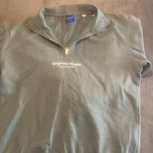 Grön tröja med dragkedja från Jack & Jones - Säljer en stilren grön tröja från Jack & Jones Originals med dragkedja och texten 'Originals Studio' på bröstet. Perfekt för en avslappnad look.