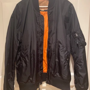 Svart bomberjacka från Alpha Industries - Säljer en klassisk svart bomberjacka från Alpha Industries med en iögonfallande orange insida. Jackan har dragkedja framtill och ribbade muddar vid ärmslut och nederkant. Perfekt för en cool och avslappnad stil. Storlek XL. Knappt använd. 