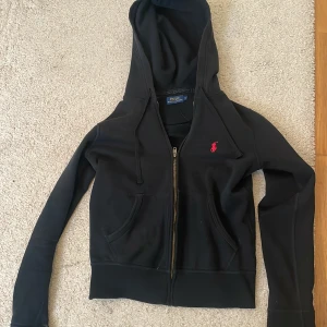 Svart hoodie från Polo Ralph Lauren - Säljer en stilren svart hoodie från Polo Ralph Lauren med dragkedja och det klassiska röda logotypbroderiet på bröstet. Perfekt för en avslappnad look. Skulle säga att den motsvarar xs.💕
