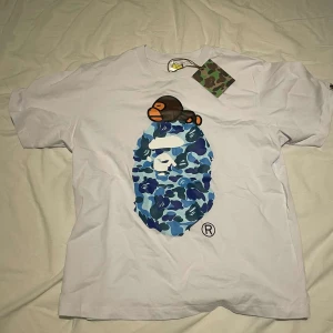 Bape t shirt - Säljer en cool vit t-shirt från A Bathing Ape med ett stort blått kamouflagemönster och en apa på framsidan. T-shirten har korta ärmar och en normal passform. Perfekt för dig som gillar streetwear!