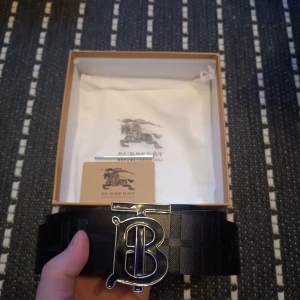 Svart bälte från Burberry - Snyggt svart bälte från Burberry med ett stort silverfärgat spänne i form av ett B. Bältet har ett diskret rutigt mönster och kommer i originalförpackning med dustbag. Perfekt för att ge en lyxig touch till din outfit. Priset kan diskuteras.