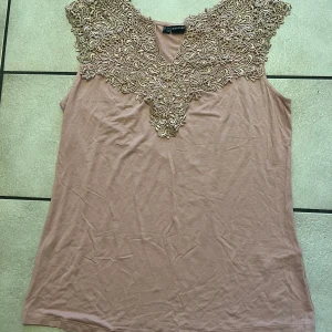 Beige ärmlös topp från Bodyflirt - Säljer en elegant beige ärmlös topp från Bodyflirt med vacker spetsdetalj vid halsringningen. Toppen är tillverkad i mjuk viskosblandning och har en avslappnad passform. Perfekt för en stilren look.