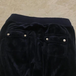 juicy couture byxor - Säljer ett par mörkblå mjukisbyxor med elastisk midja och snörning. Byxorna har två fickor fram och två bak med knappdetaljer. Perfekta för en avslappnad stil.