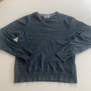 Blå tröja från Hilfiger Denim - Säljer en stilren blå tröja från Hilfiger Denim. Tröjan har en liten broderad logga på bröstet och är tillverkad i ett mjukt material. Har en liten defekt vid lappen där det vi är lite trådar men annars finns det inga andra defekter. 