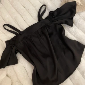 Svart offshoulder topp - Snygg svart offshoulder topp med smala axelband och korta ärmar. Toppen har en luftig passform och är perfekt för en stilren look. Materialet ger en elegant känsla och passar bra till både jeans och kjol.