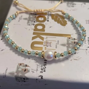 armband med söttvattenspärla - Snyggt armband med ljusblå facetterade pärlor, guldfärgade detaljer och en större vit sötvattenspärla i mitten. Justerbar beige snörning gör det enkelt att anpassa storleken. Perfekt för dig som gillar stilrena och trendiga accessoarer.