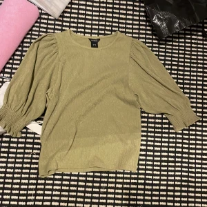 Grön blus med puffärm från Lindex - Snygg ljusgrön blus från Lindex med puffiga trekvartsärmar och smockdetaljer vid ärmsluten. Blusen har en lätt strukturerad yta och rak passform, perfekt för dig som gillar en trendig och avslappnad stil.