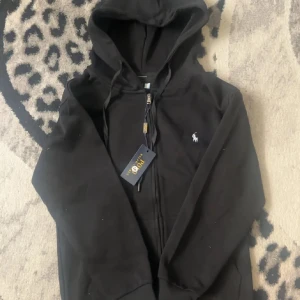 Svart hoodie från Polo Ralph Lauren - Svart hoodie från Polo Ralph Lauren med vit broderad logga på bröstet. Tröjan har dragkedja, huva med snörning och klassisk slim fit. Perfekt för dig som gillar stilren och enkel streetwear.
