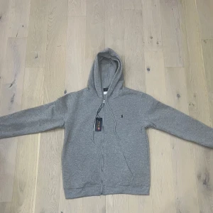 Grå hoodie från Polo Ralph Lauren - Grå hoodie från Polo Ralph Lauren med dragkedja framtill, huva med snörning och klassisk logga på bröstet. Tillverkad i mjuk bomullsmix och har långa ärmar samt två fickor framtill. Perfekt för en chill och avslappnad stil.