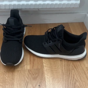 Svarta Adidas Ultraboost sneakers - Svarta Adidas Ultraboost sneakers med stickad ovandel och klassiska tre ränder på sidan. Skorna har vit Boost-sula för extra komfort och svart yttersula. Perfekta för dig som gillar stilrena och sportiga sneakers med modern känsla.