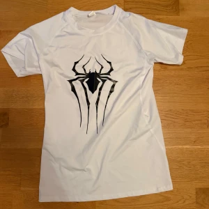 Vit t-shirt med spindeltryck compression - Snygg vit t-shirt med ett stort svart spindeltryck på bröstet, inspirerat av Spiderman. T-shirten har korta ärmar och rund hals. Materialet känns lätt och sportigt, perfekt för dig som gillar coola prints och vill sticka ut.
