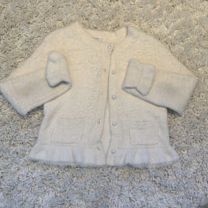 Vit kofta från Lindex med pärlknappar - Säljer en supermjuk och fluffig vit kofta från Lindex med pärlknappar framtill och två små fickor. Koftan har lång ärm och är tillverkad i en mix av akryl och polyester. Koftan har volangkant nertill och kostar runt 299kr! Köparen står för priset på frakten. Priset går att diskuteras och om man vill ha mer bilder så är det bara att kontakta mig här på plick💗💐