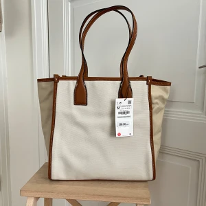 Beige shoppingväska från Zara - Stilren shoppingväska från Zara i beige bomullstyg med bruna skinnhandtag och detaljer. Väskan har rejäla handtag, rymligt fack och innerficka med dragkedja. Perfekt för att bära allt du behöver till skolan eller stan.