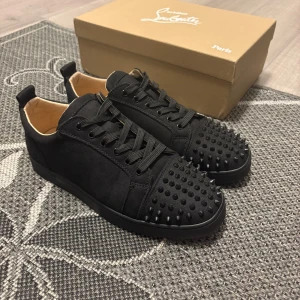 Svarta Christian Louboutin sneakers med nitar - Säljer ett par svarta sneakers från Christian Louboutin med coola svarta nitar på tån och klassisk röd sula. Skorna har snörning, rund tå och är tillverkade i mocka med läderfoder. Perfekt för dig som vill sticka ut med en lyxig och edgy stil.