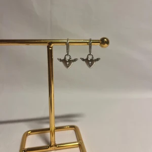 Silverörhängen med hjärtan och vingar - Snygga örhängen i silverfärg med hängande hjärtan som har små vingar på sidorna och ett nyckelhål i mitten. Ringen är dekorerad med små stenar för extra bling. Perfekta för dig som gillar unika detaljer och cool design.