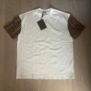 Burberry t-shirt med rutiga ärmar - Snygg vit Burberry t-shirt med klassiska rutiga ärmar i brunt, svart, vitt och rött. T-shirten har rund hals och är kortärmad. Materialet är mjuk bomull och passformen är normal. Perfekt för dig som gillar stilrena designer med en twist.