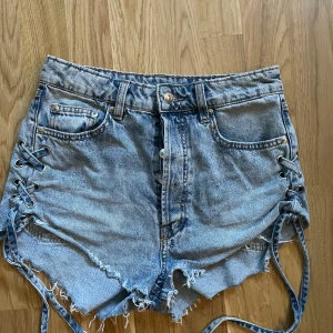 Blå jeansshorts med snörning och hjärta - Snygga blå jeansshorts med hög midja och råa kanter. På sidorna finns cool snörningsdetalj och på baksidan är en broderad hjärtformad patch. Klassisk femficksmodell i denim, perfekt för sommarens alla äventyr.