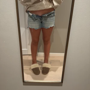 Blå jeansshorts med broderade fickor - Snygga ljusblå jeansshorts från Miss Me med slitna detaljer och råa kanter. Baksidan har broderade fickor med paisleymönster och silverfärgade nitar. Klassisk femficksmodell och knappstängning framtill. Perfekta för sommarens chill dagar.