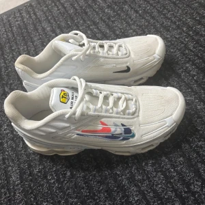 Nike Air Max Plus vita sneakers - Nike Air Max Plus sneakers i vitt med coola detaljer i rött och blått på sidan. Skorna har snörning, mesh och syntetmaterial, samt den klassiska Tn-loggan på plösen. Snygg och sportig vibe med synlig Air-sula och svart mini-Swoosh.