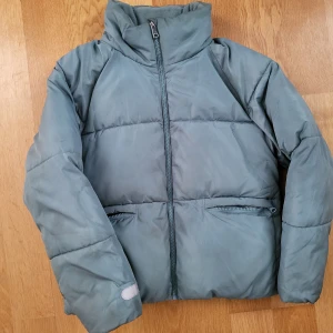 Turkosgrå pufferjacka från Lindex, strl 158 - Snygg turkosgrå pufferjacka från Lindex i storlek 158. Jackan har hög krage, dragkedja framtill och två fickor med dragkedja. Vattenavvisande material och reflekterande detalj på ärmen. Perfekt för kyliga dagar när du vill hålla dig varm och stilren.