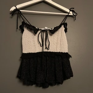 Stickad topp med volang från Zara - Supersöt stickad topp från Zara i vitt och svart med volangkant och knytband vid axlarna. Toppen har en ribbad överdel, spetsdetaljer och en glittrig, svart volang nedtill. Perfekt för dig som gillar unika och trendiga plagg med extra detaljer.