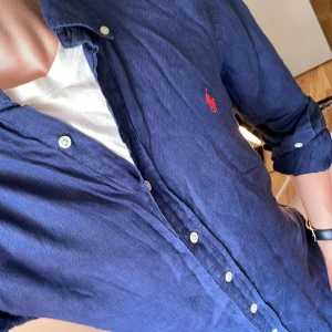 Polo Ralph Lauren Linneskjorta  - Säljer en marinblå långärmad skjorta från Polo Ralph Lauren i slim fit. Skjortan är i linne och har klassisk krage, vita knappar och den ikoniska röda broderade loggan på bröstet. Perfekt för dig som gillar stilrena och snygga plagg.                           Plagget har några små små imperfektioner (fråga om bild). Men i övrigt bra skick                                                              Osäker på priset så kom med förslag!!!!!